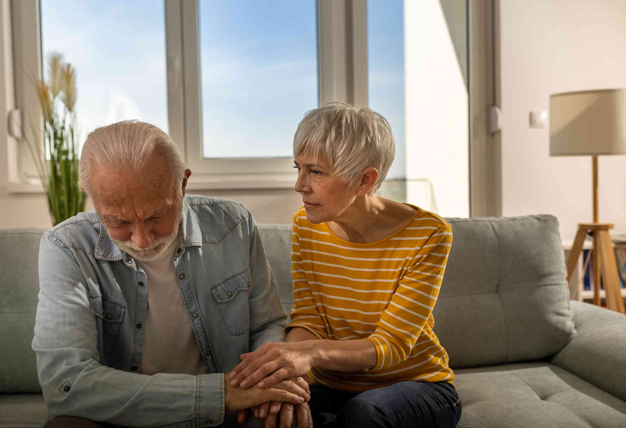 A couple is seated on a couch. A man looks sad while his wife holds his hand, offering comfort.